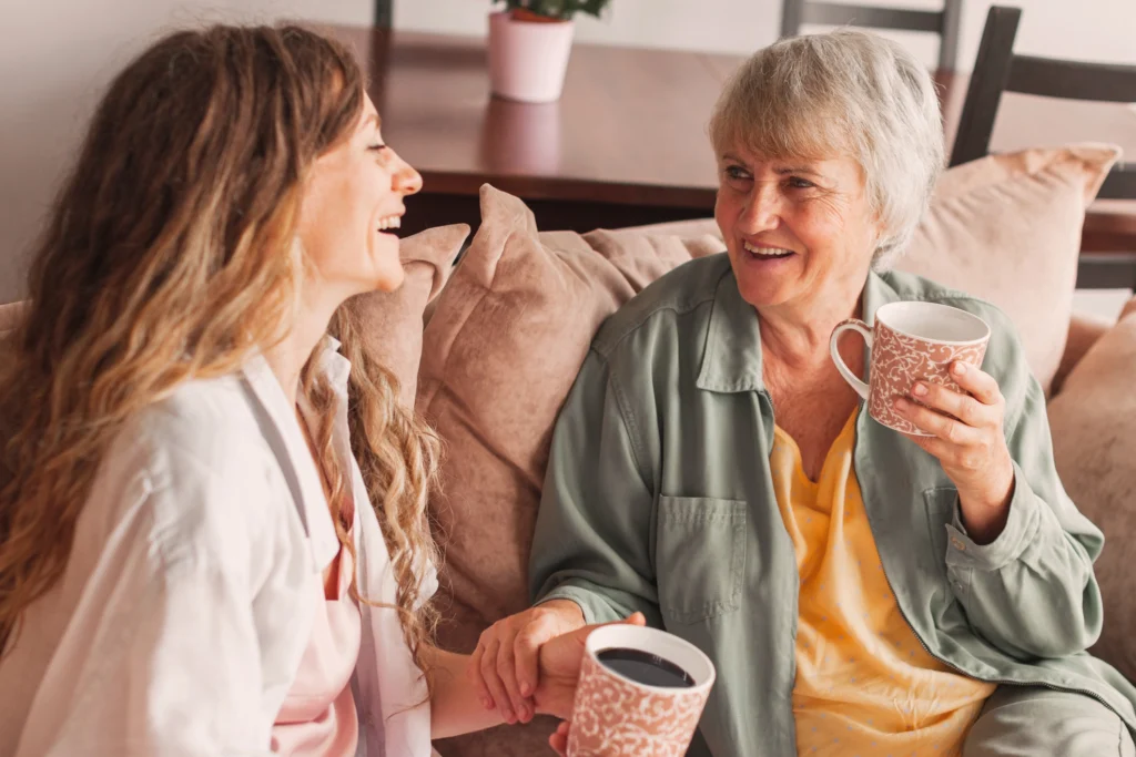 An adult daughter and her elderly mother sitting together on a sofa, laughing and holding mugs. This image relates to: 20 things not to say to someone with dementia.