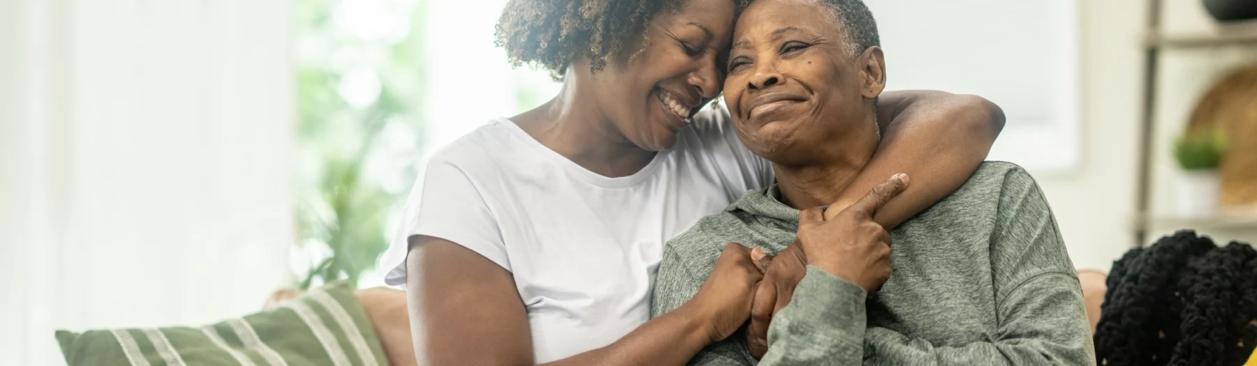 An older mother and her adult daughter, sitting on a couch embracing and holding hands. This image relates to: 20 things not to say to someone with dementia.