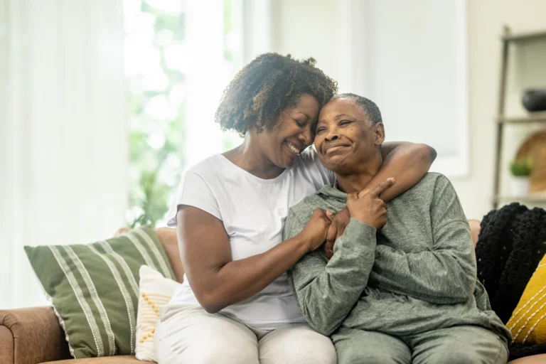 An older mother and her adult daughter, sitting on a couch embracing and holding hands. This image relates to: 20 things not to say to someone with dementia.